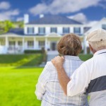 Senior couple standing in the foreground staring at a two story white colonial house with front porch. The man on the right has his left hand on the woman's shoulder.
