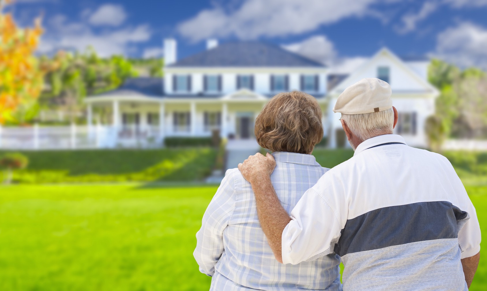 Senior couple standing in the foreground staring at a two story white colonial house with front porch. The man on the right has his left hand on the woman's shoulder.