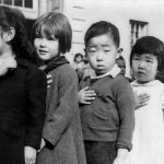 Photo Credit: Children at the Weill public school in San Francisco pledge allegiance to the American flag in April 1942, prior to the internment of Japanese Americans. Photo attributed to Dorothea Lange(w). Public Domain.