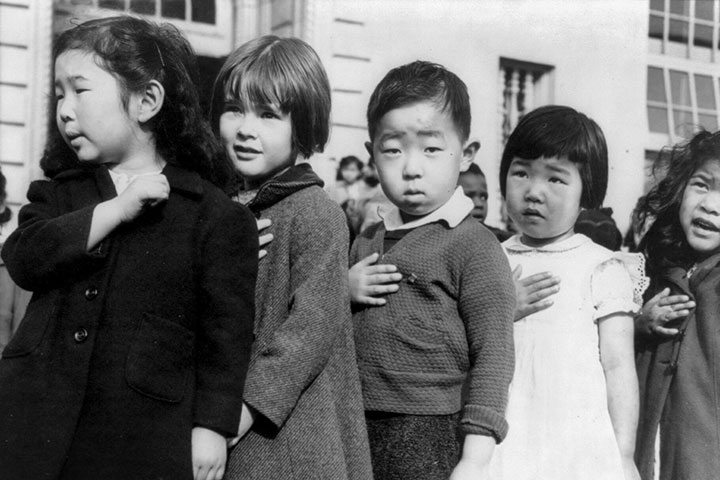 Photo Credit: Children at the Weill public school in San Francisco pledge allegiance to the American flag in April 1942, prior to the internment of Japanese Americans. Photo attributed to Dorothea Lange(w). Public Domain.