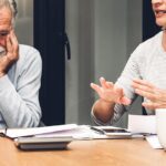 An older couple sits at a table discussing financial documents. The woman is speaking and the man has his heads in his hands with a worried look on his face.