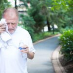 Older man out walking in the park. He is holding a water bottle in his left hand and toweling his face with his right hand. It appears to be a very hot day.