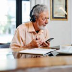 Senior male wearing headphones and holding a cellphone. He is seated at a table and looking at an open laptop computer.