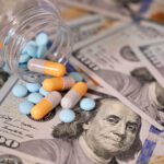 A table covered in one hundred dollar bills, a small clear bottle is lying on it's sides with various pills spilling out of it and onto the money.
