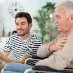Older father and adult son seated. Father is in wheelchair. Son is on couch. Son showing father something on a tablet.