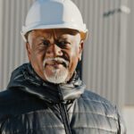 Older African-American gentleman dressed in a clack winter jacket wearing a white hard hat.