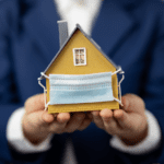 Person holding a model of a yellow house in outstretched hands. There is a surgical mask on the front of the house covering the door. Image representative of senior housing during th pandemic.