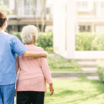 rear view of a nurse helping an elderly patient walking on an outdoor path at an assisted living facility